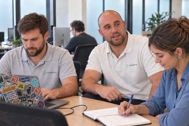Guillermo working on a sticker-covered MacBook, Agustín speaking to someone outside the frame and a colleague taking notes in an office scene.